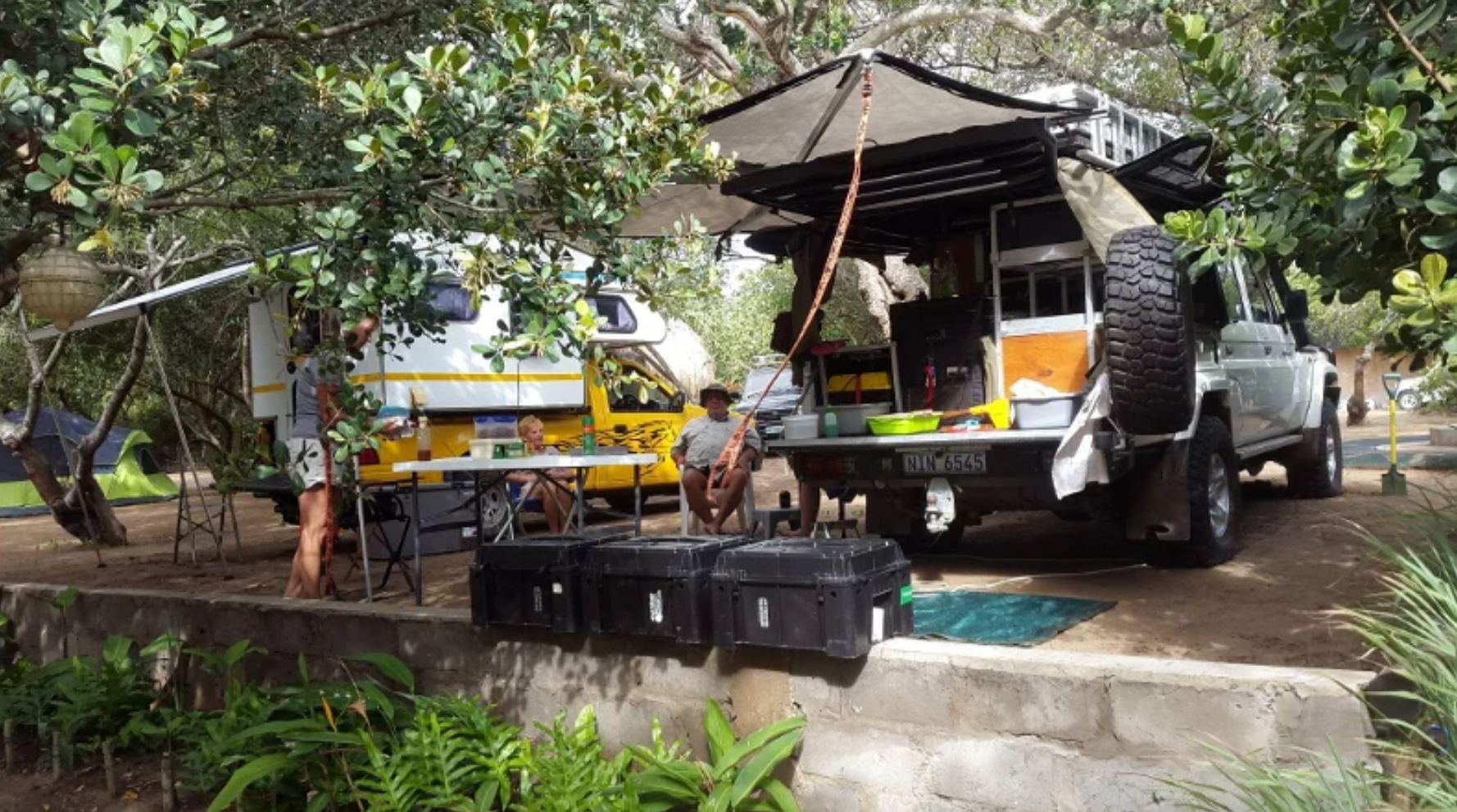 Guests relaxing at a camp table beside fully kitted overland vehicles