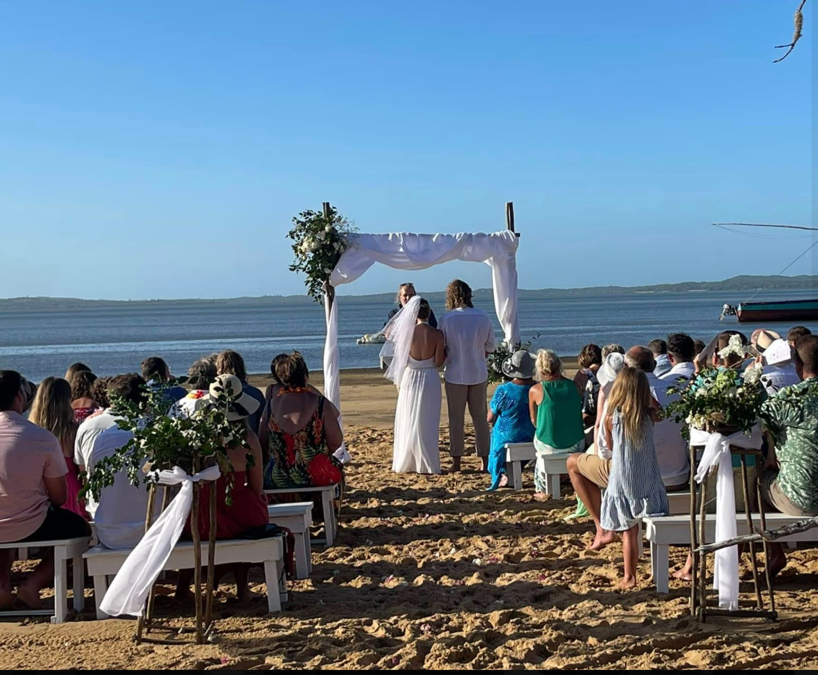 A barefoot wedding ceremony on the beach at Bemugi's with guests seated beneath a floral arch