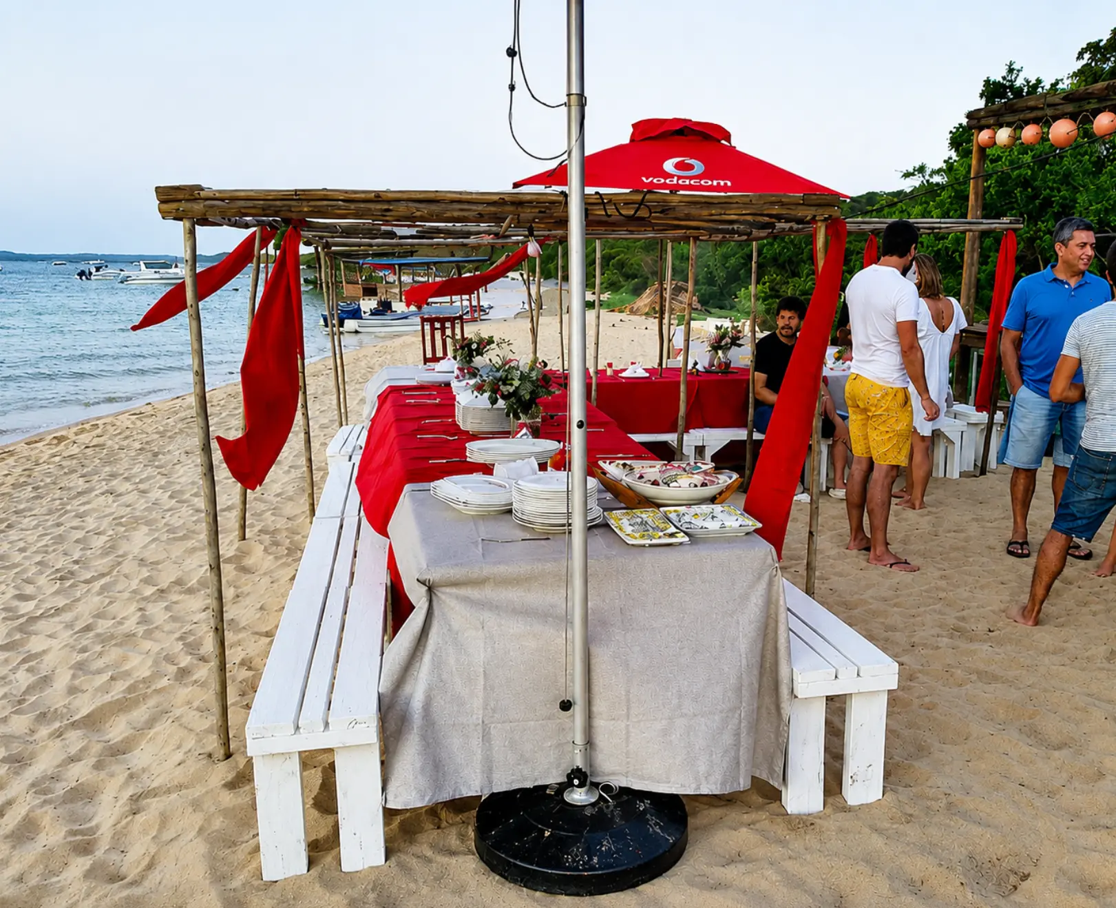 A relaxed beachside dining setup with tables, benches and guests by the water