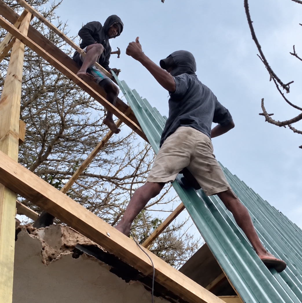Two team members rebuilding the roof at Bemugi's Place, one giving a thumbs up
